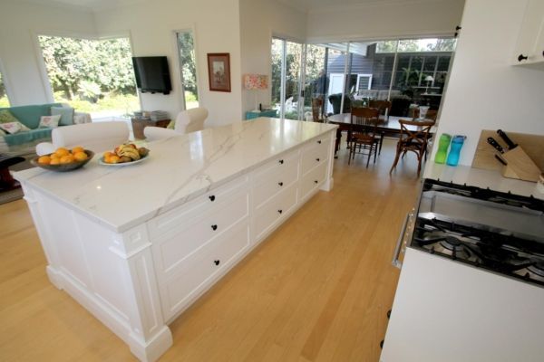 White kitchen island with marble countertop, drawers, and fruit bowl; adjacent to hardwood floors and a dining area — PK Kitchen Design in West Ipswich, QLD