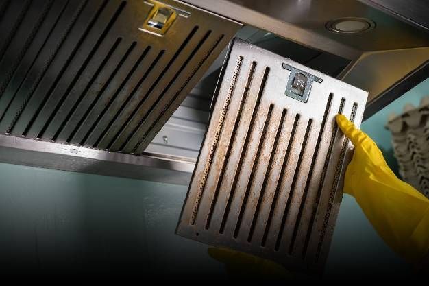 Person in yellow gloves removes a dirty metal range hood filter from a stainless steel hood.