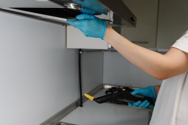 Person in blue gloves cleaning a kitchen range hood with a steam cleaner.