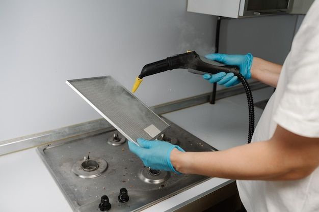 Person cleaning a metal kitchen filter over a stovetop with a steam cleaner, wearing blue gloves.