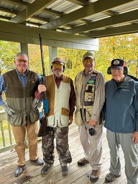 Four men, wearing vests and ear protection, stand outdoors with a shotgun, likely at a shooting range.