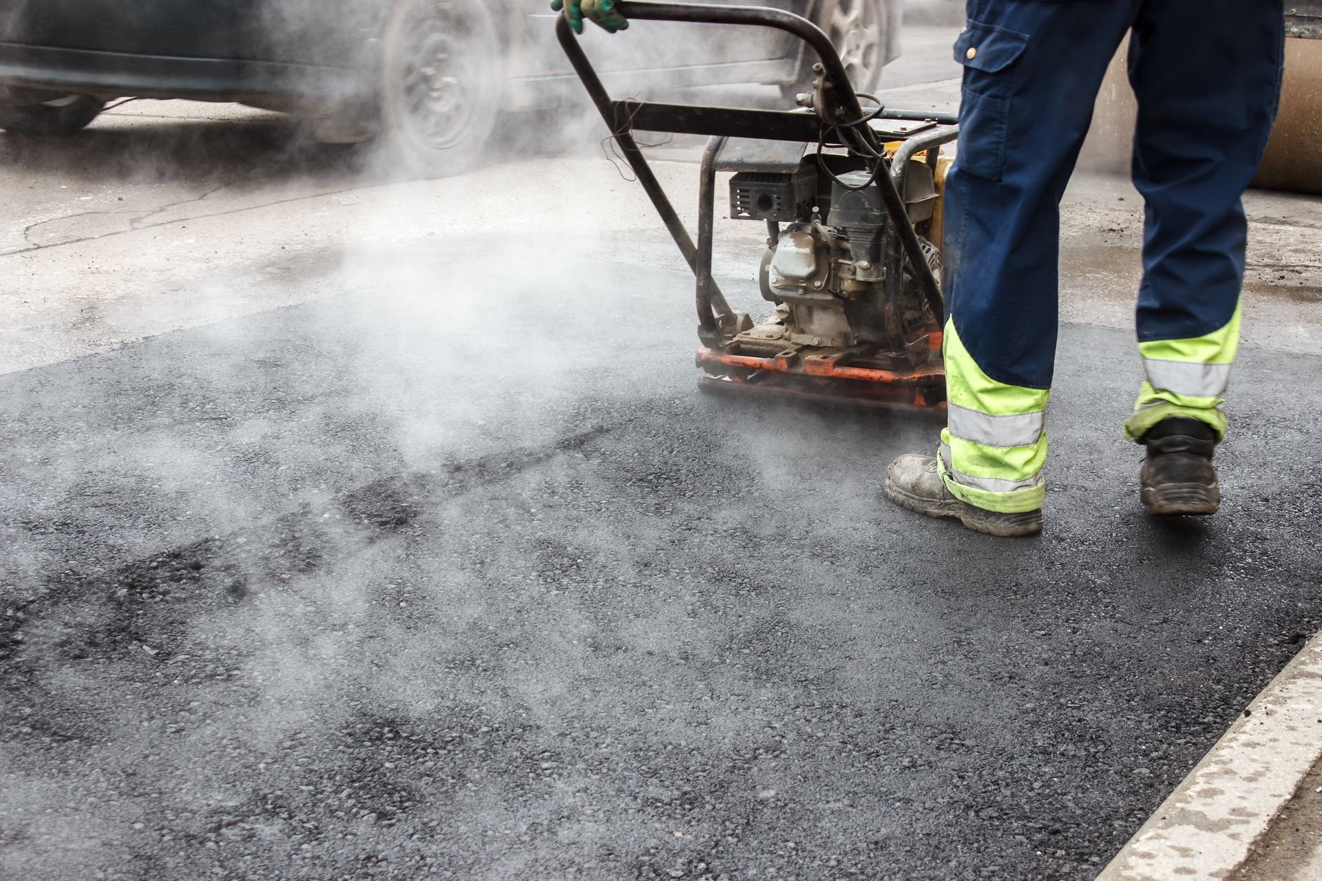 Person compacting fresh asphalt with a vibrating plate compactor on a road. Steam rises from the surface.