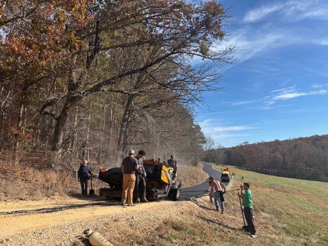 Road crew working on a gravel road, with a machine spraying materials. Blue sky, autumn trees.