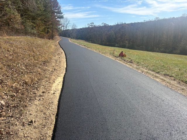 A newly paved asphalt path curves through a grassy field and treeline under a blue sky.