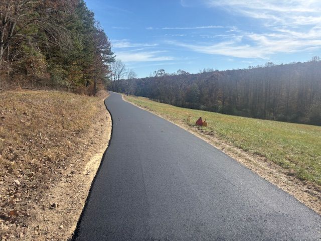 Paved asphalt path winding through a grassy field and treeline under a blue sky.