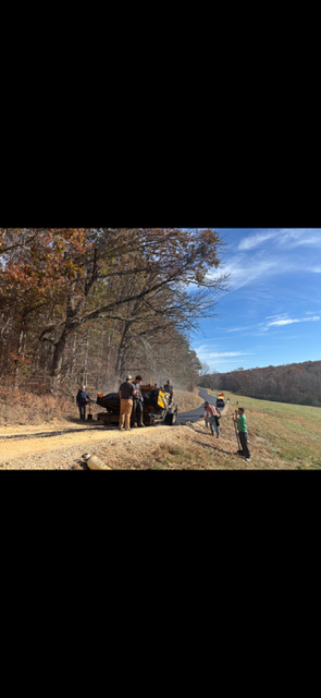 People standing near a vehicle on a dirt road next to a field. Trees line the road on one side, blue sky.