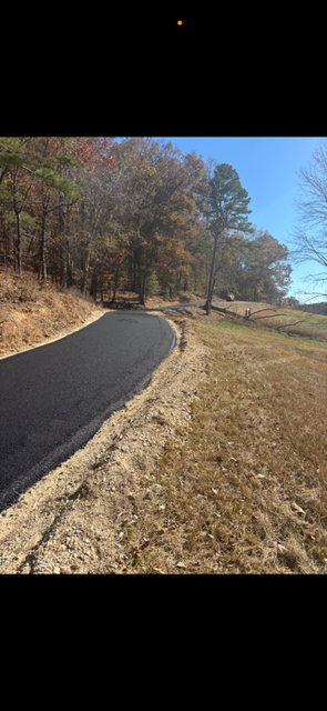 Newly paved road curves through a rural landscape, flanked by trees and dry grass under a blue sky.