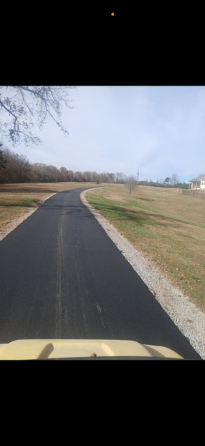 An asphalt road curves through a grassy field, trees in the background, a house on the right.