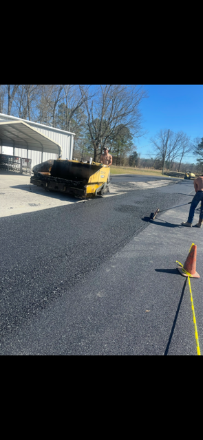 Newly paved asphalt driveway leading toward a house, bordered by grass and a chain-link fence.