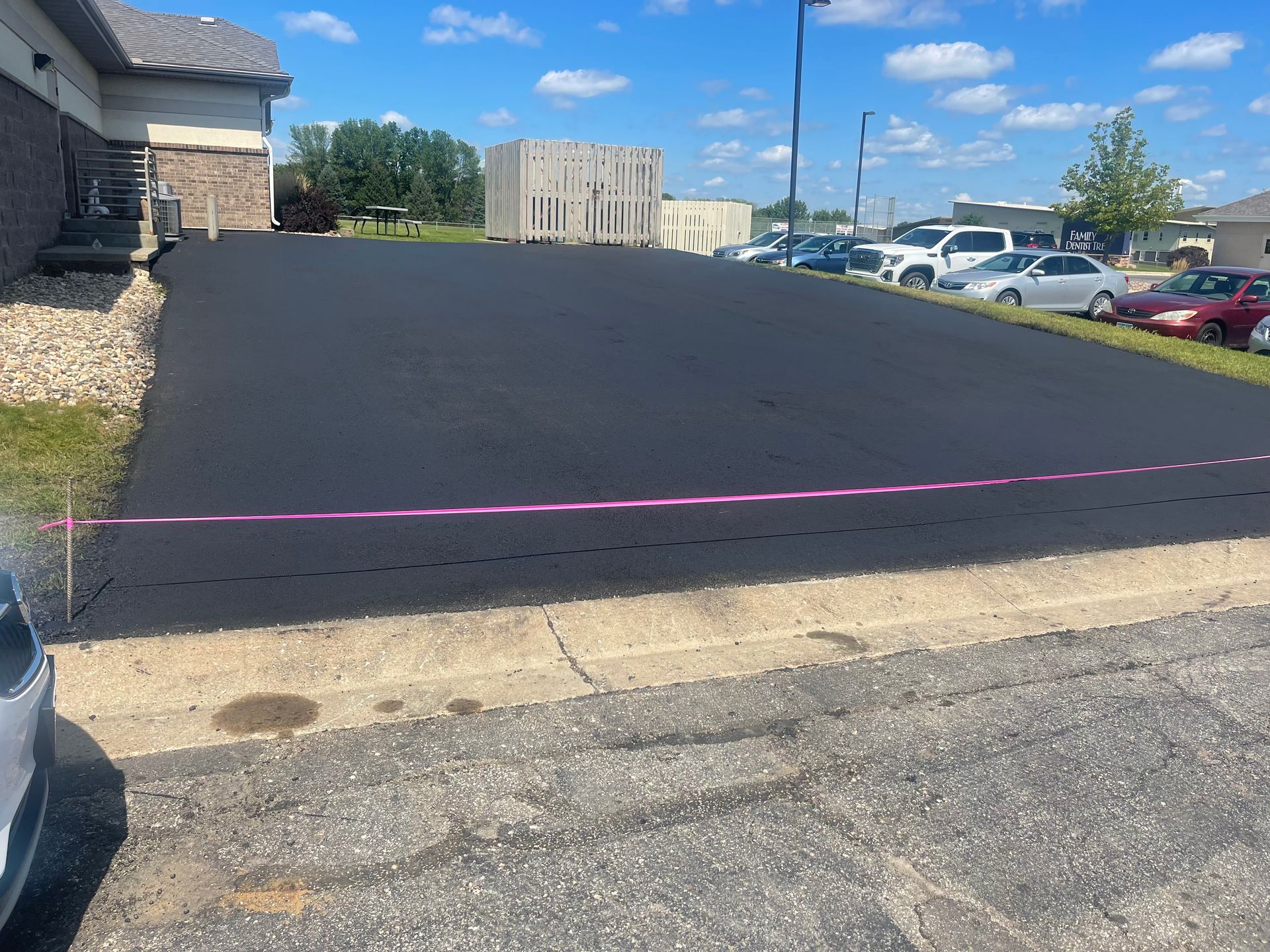 Newly paved asphalt parking lot with pink caution tape, parked cars in background.