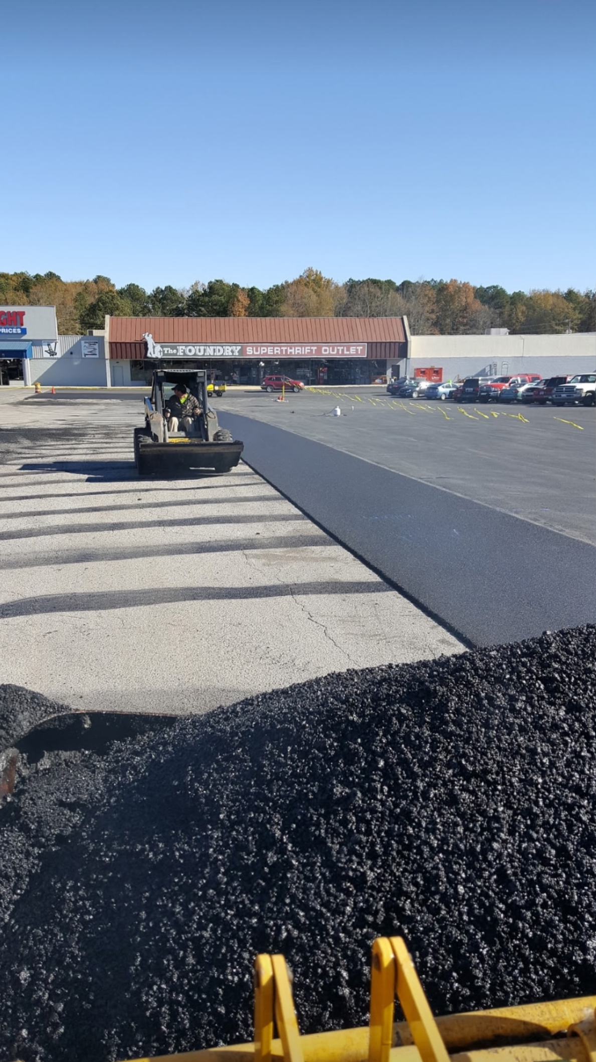 Asphalt paving a parking lot; a steamroller compacts the fresh blacktop near a business building under a clear sky.