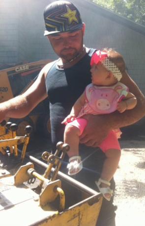 Man holding a baby near construction equipment. The baby is wearing a pink outfit and headband. The man is wearing a hat.
