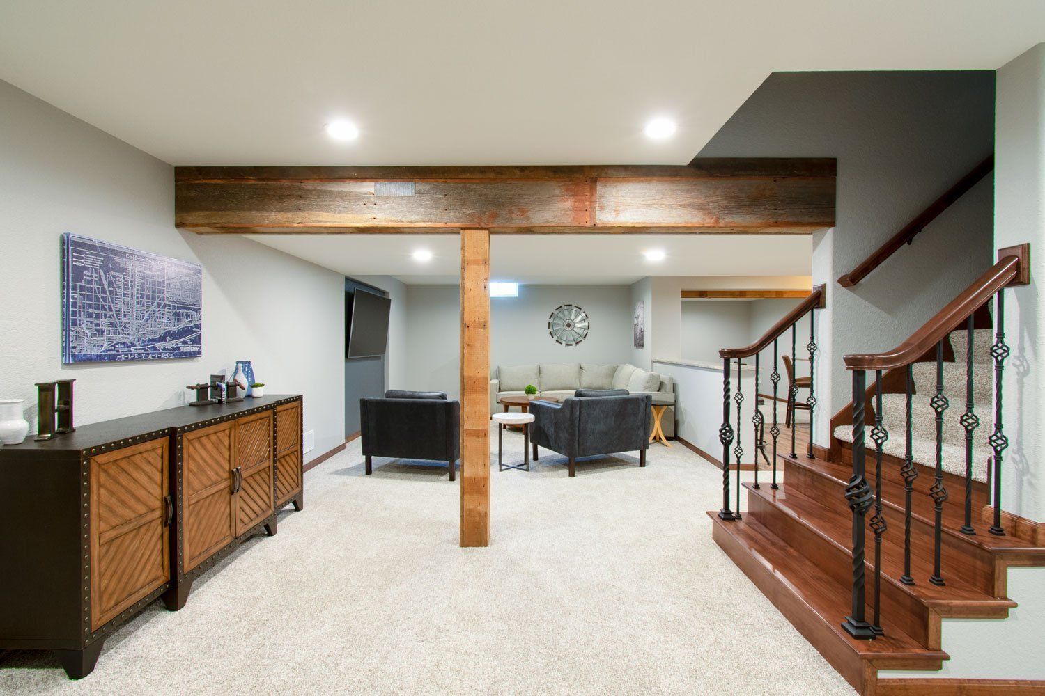 Finished basement living area with wood stairs, exposed beam, and dark furniture amid light carpet.