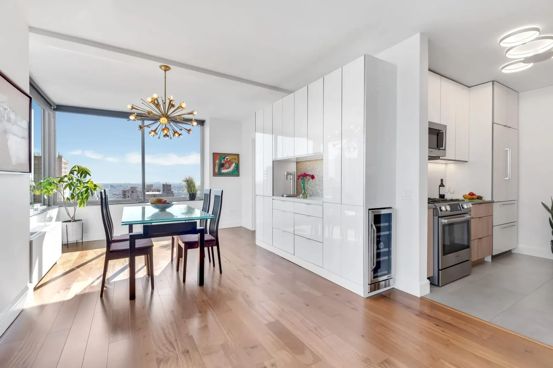 Bright open dining and kitchen area with wood floors, white cabinets, and large windows overlooking water.
