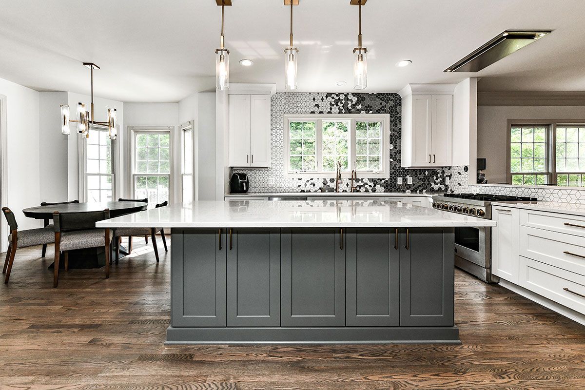 Bright modern kitchen with gray island, white cabinets, wood floors, and dining area by large windows.