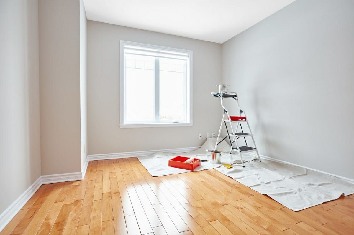 Bright empty room with hardwood floor, window, and painting supplies on a drop cloth