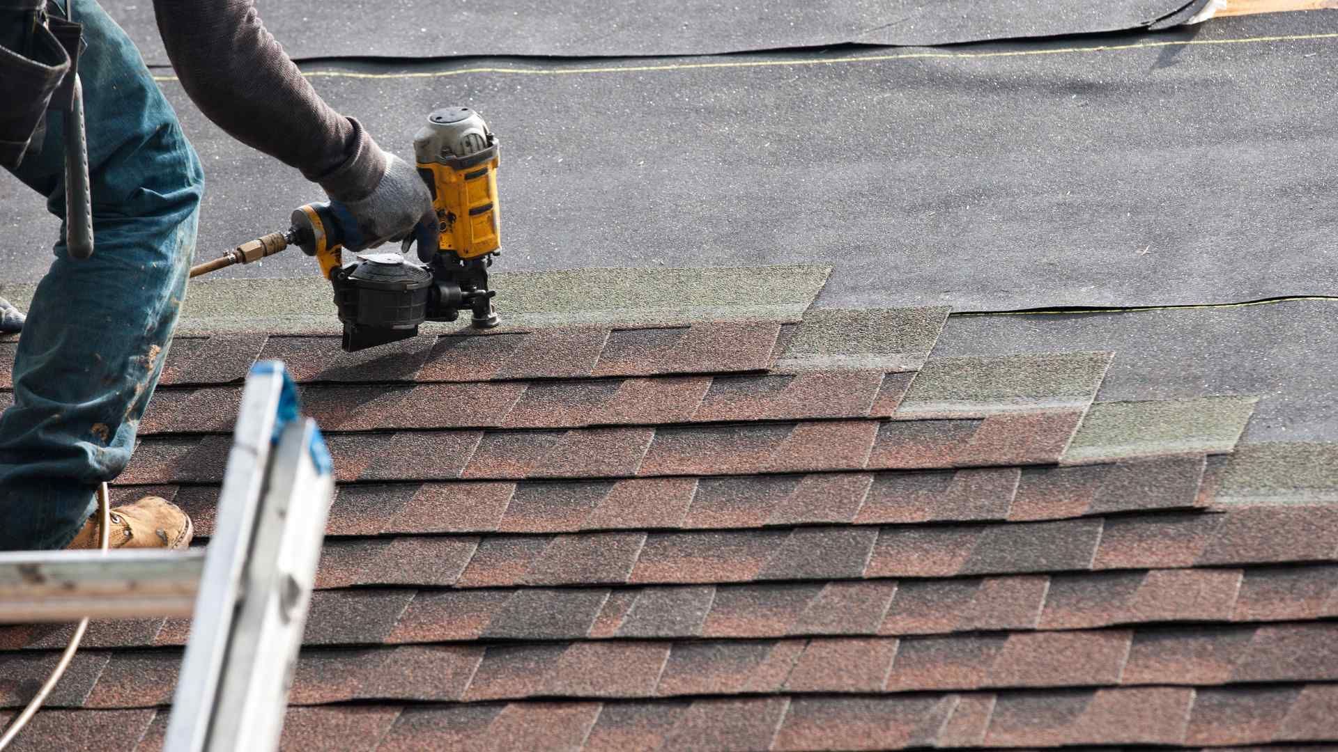 Worker using a nail gun installing dark roof shingles on a sloped rooftop
