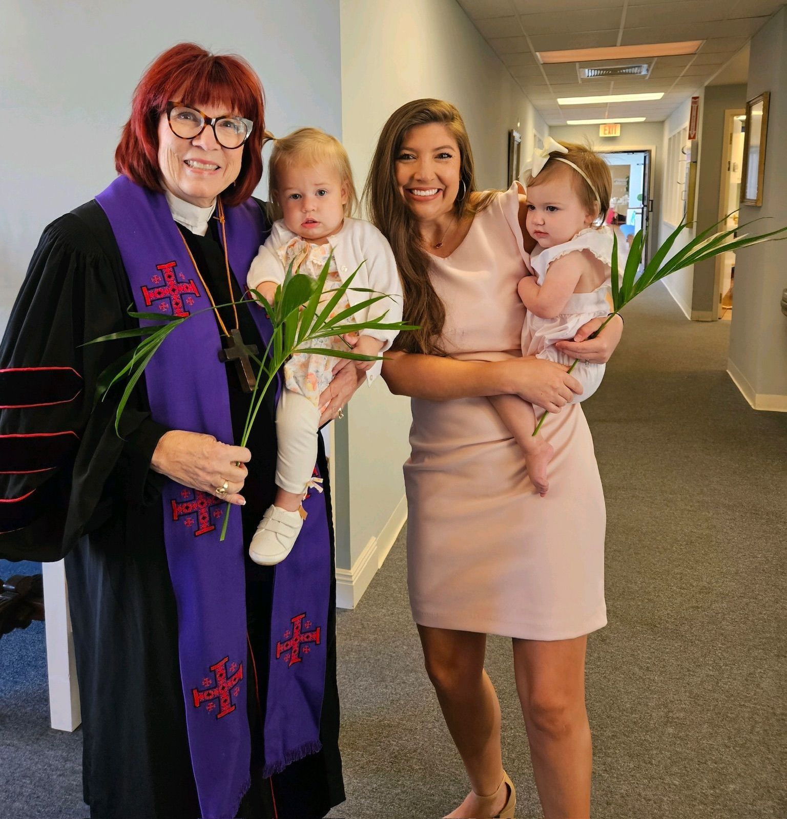 Woman in clergy robes, young woman, and two babies holding palm fronds in a hallway.