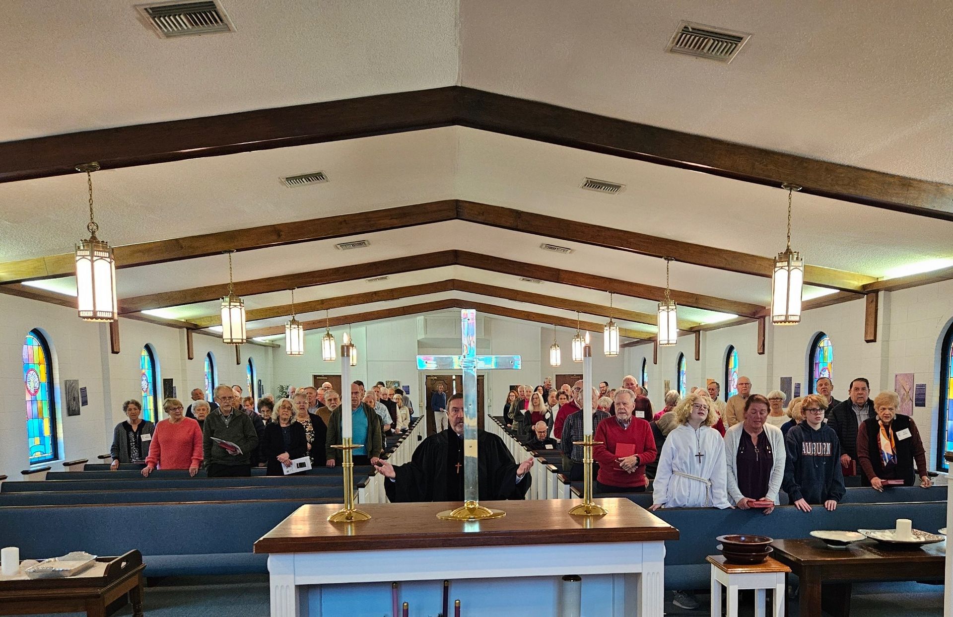 People standing inside a church, facing an altar with a cross. Sunlight streams through windows.