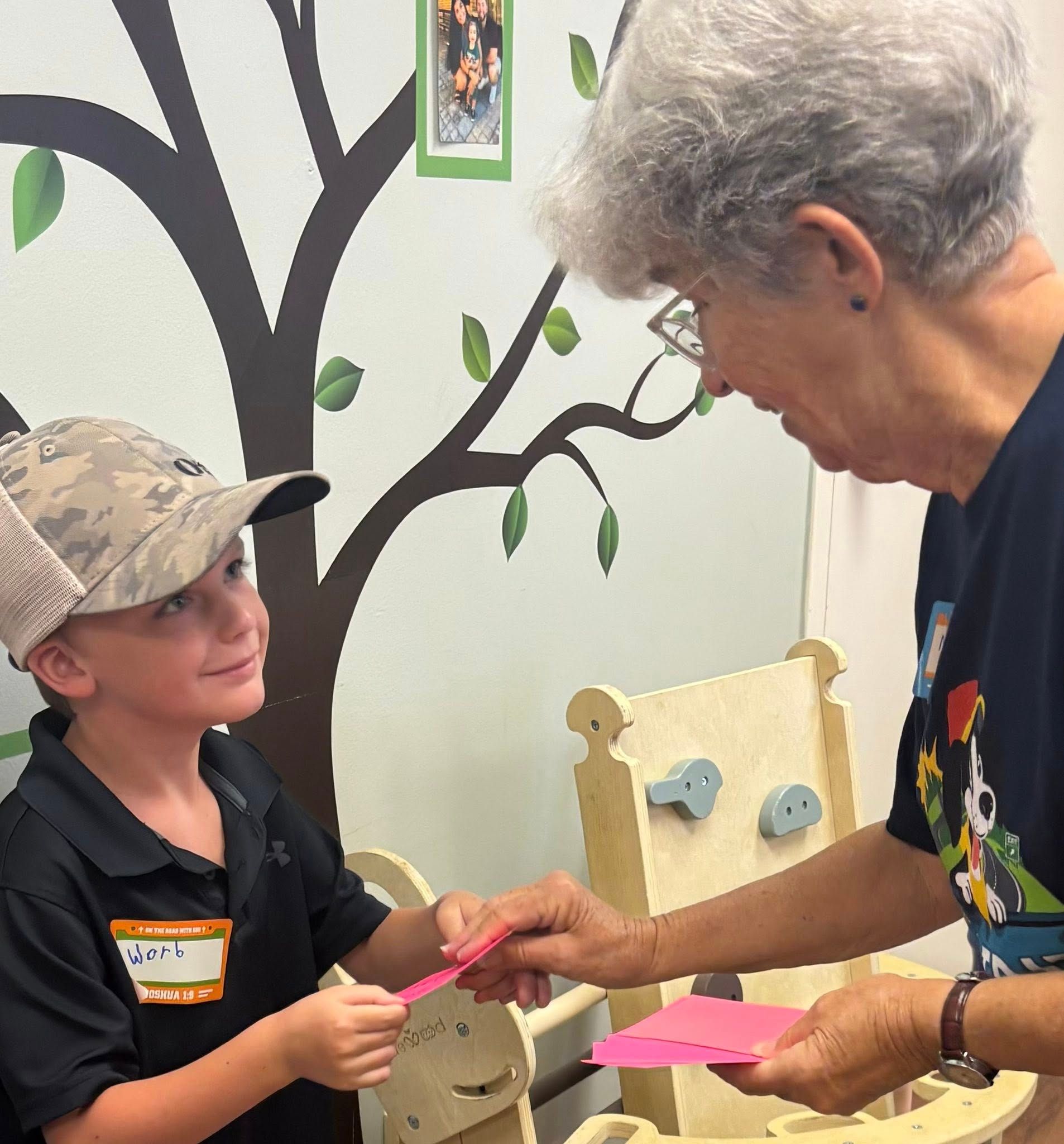 Boy in camo hat handing pink card to older woman in front of a tree mural.