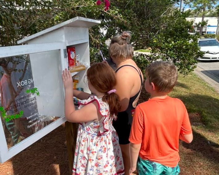 Children and a woman at a small outdoor food pantry, reaching inside. White cabinet with glass door, sunny day.