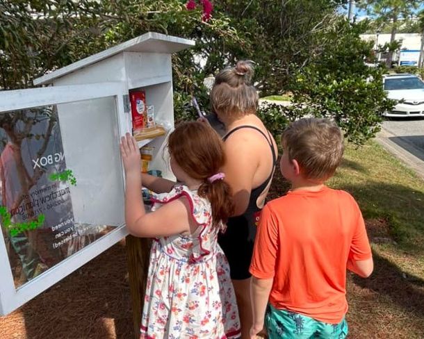 Children and woman at a small food pantry box. One child reaches inside; sunny outdoor setting.