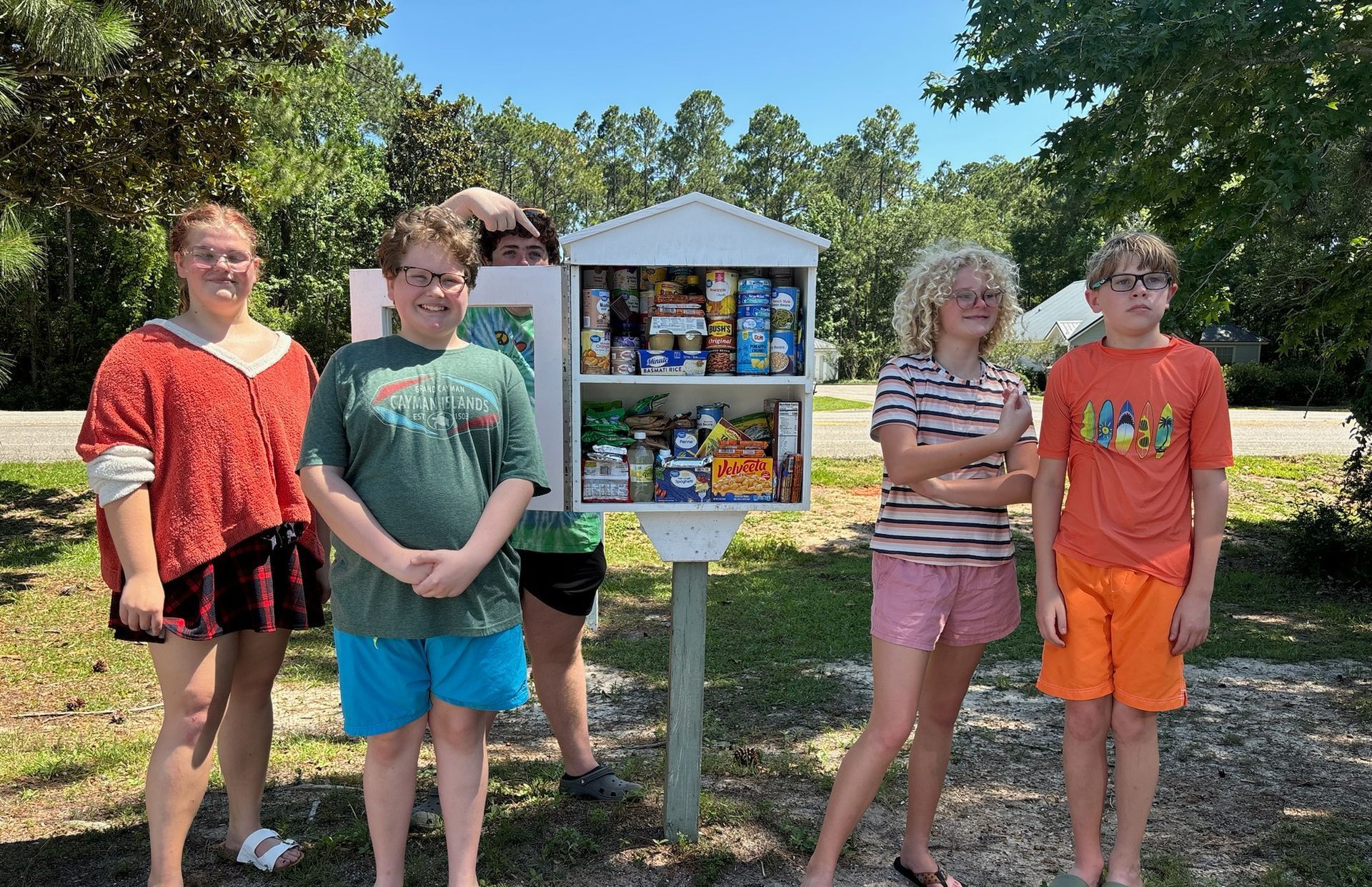Five kids pose near a white food pantry outdoors. Some are smiling, with snacks visible.