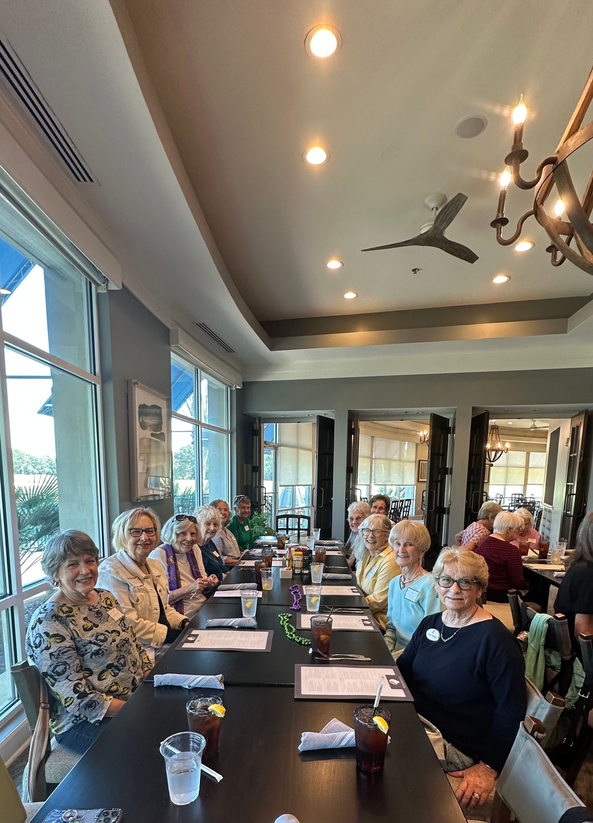 A group of older women sit at a long table in a restaurant, smiling at the camera.