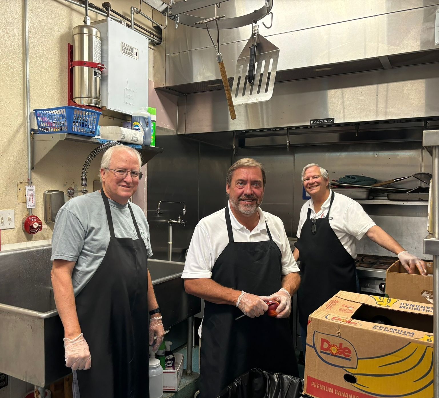 Three men in a commercial kitchen, wearing aprons and gloves, prepping food near stainless steel sinks and shelves.