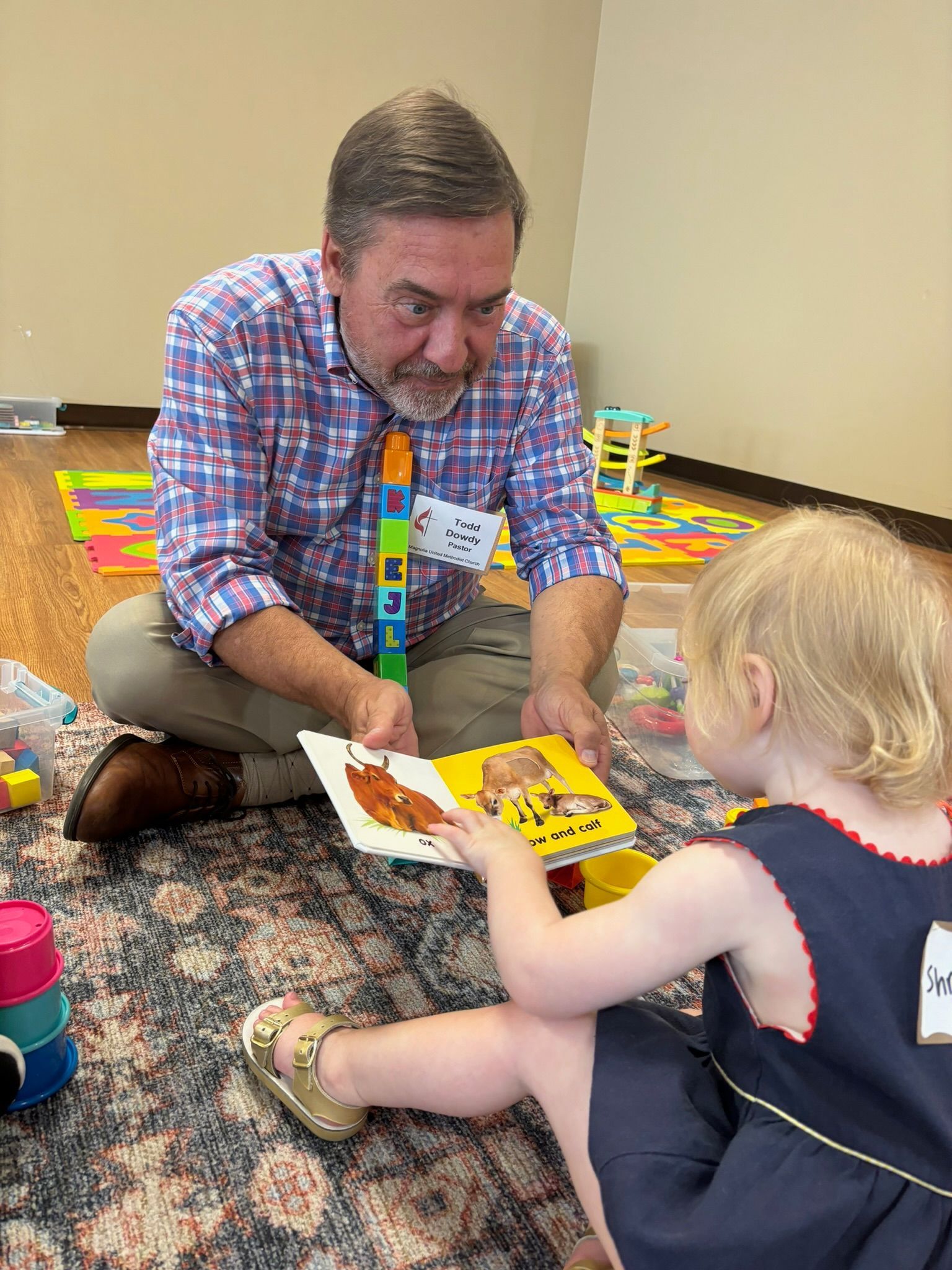 Man in plaid shirt reads a book to a young girl in a room with toys; both are sitting on the floor.