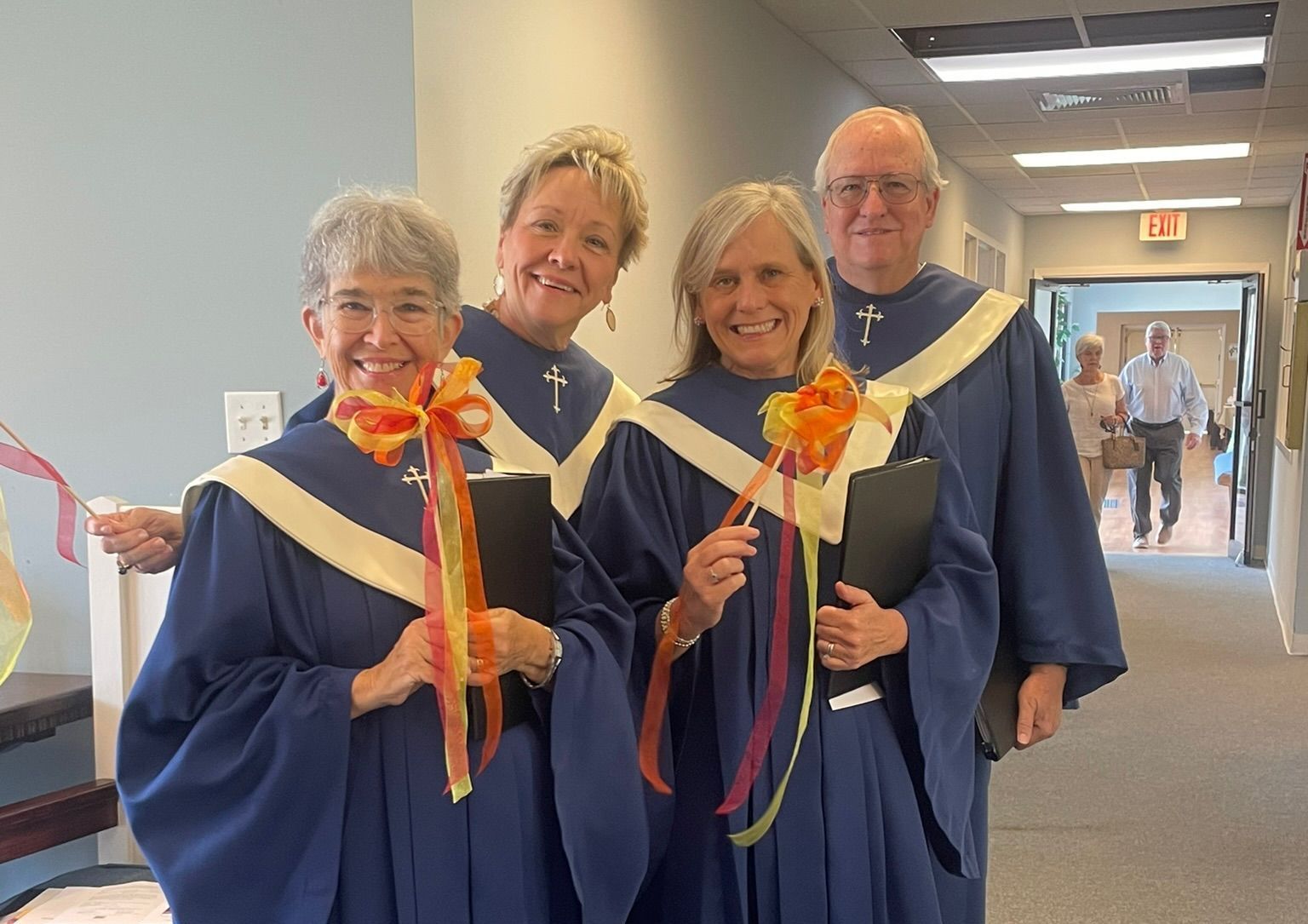 Choir members in blue robes with ribbons and smiles in a hallway, one man and three women.