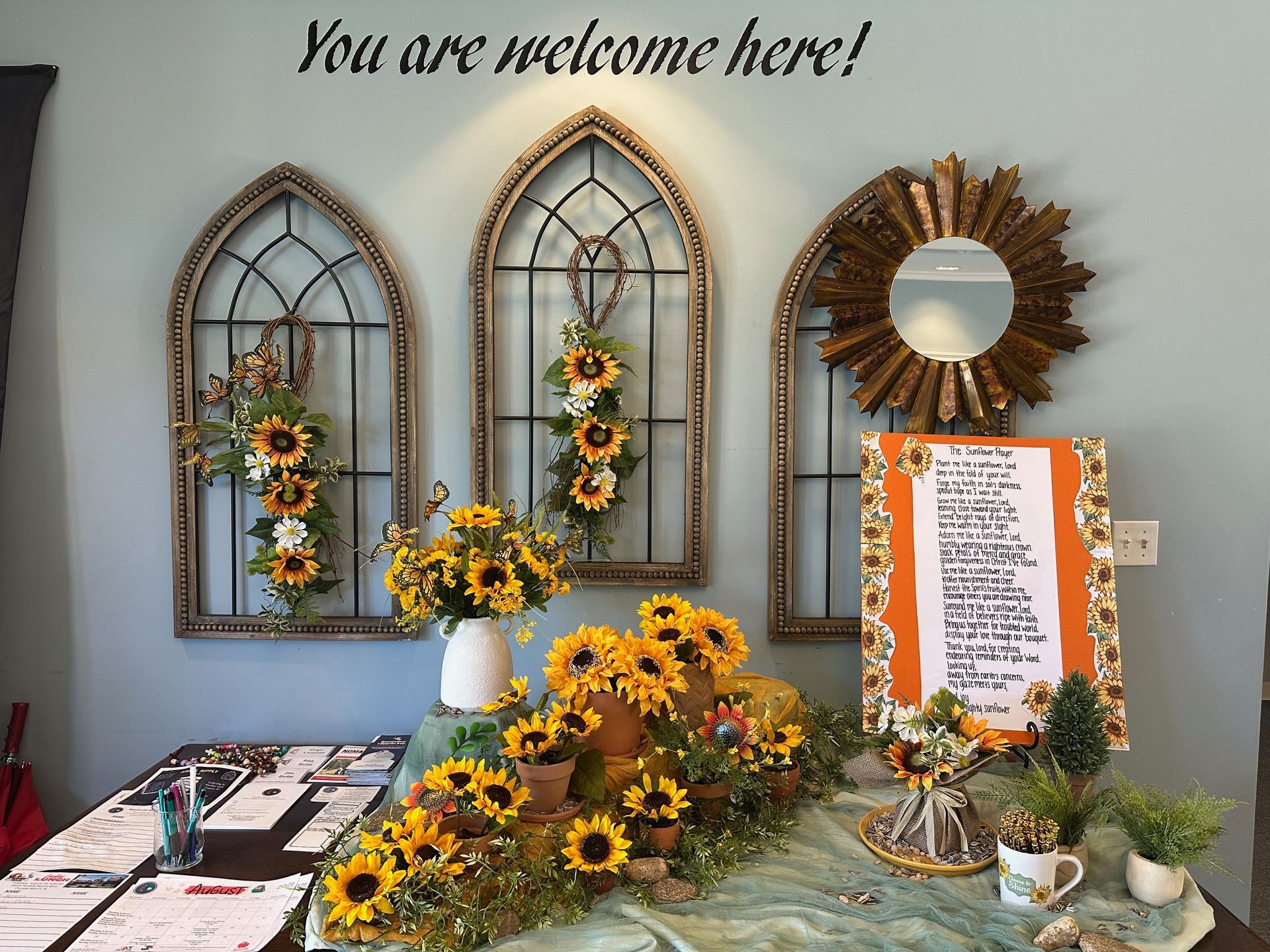 Sunflowers and decor welcome visitors. Three arched window frames, mirror, and sign adorn a teal wall.