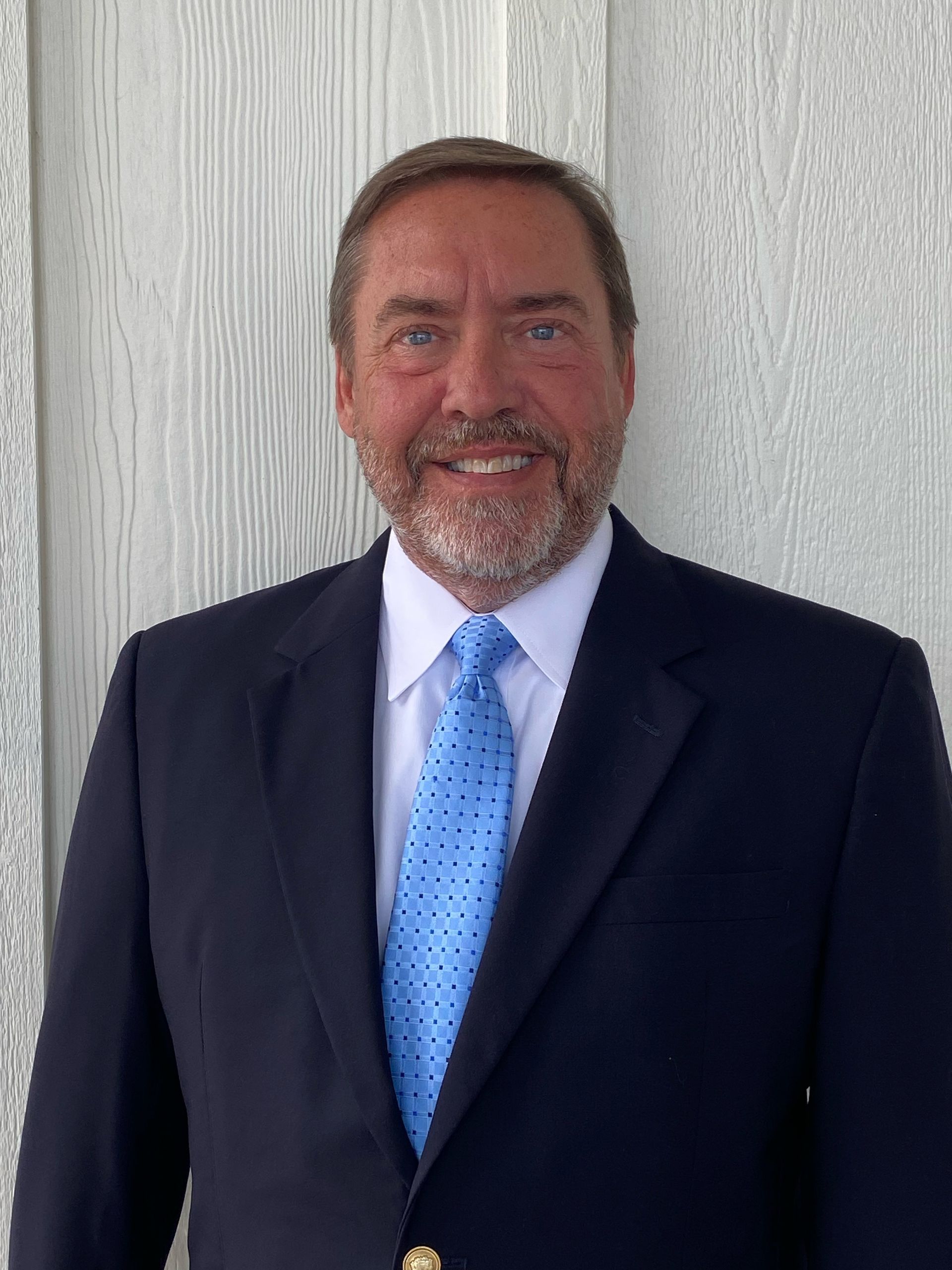 Man in a navy blazer and blue tie smiles in front of a white wall.