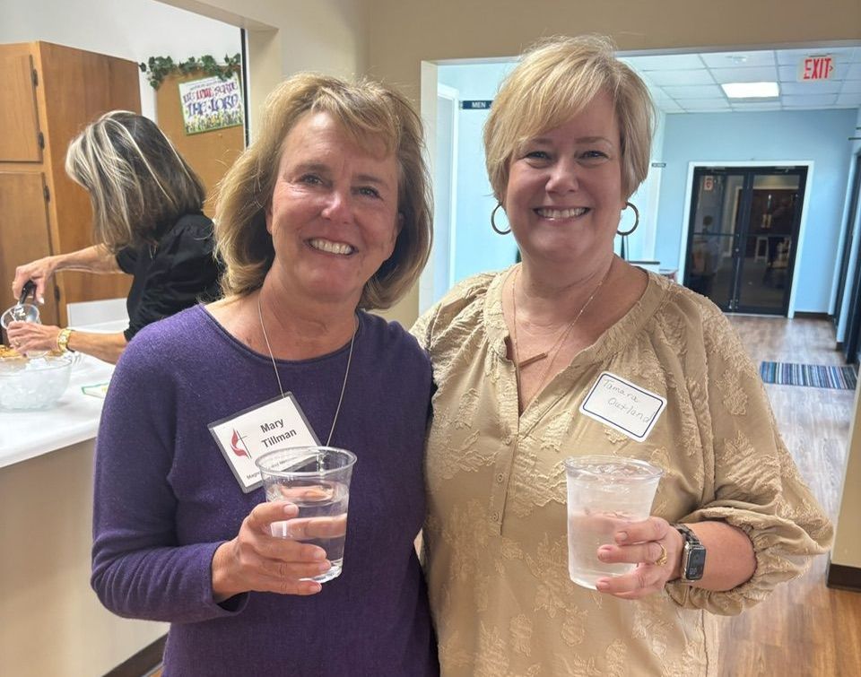 Two women smiling, holding drinks at an event. One wears purple, the other tan. Bright, indoor setting.