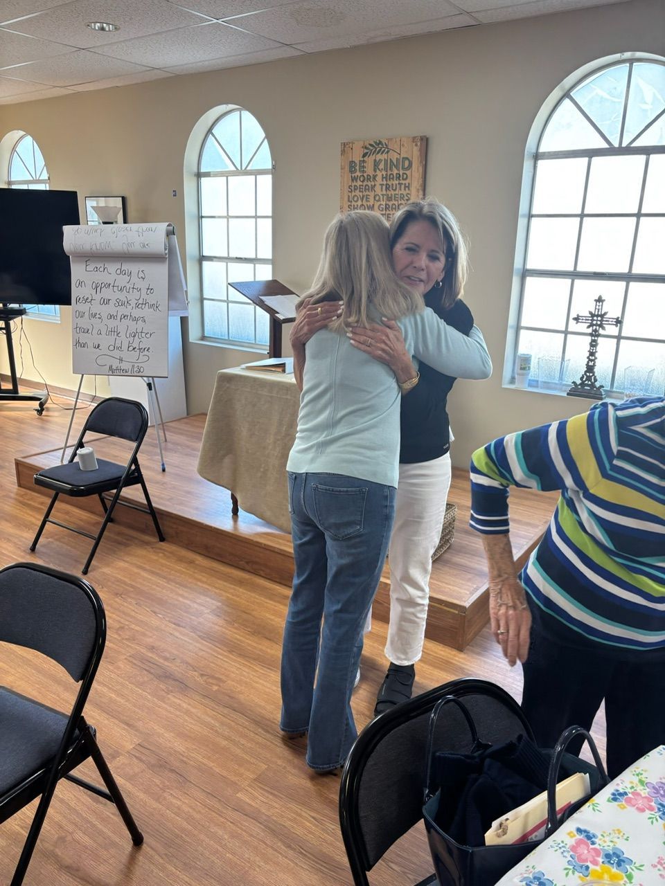 Two women hugging inside a room with arched windows and a pulpit. Another woman watches.