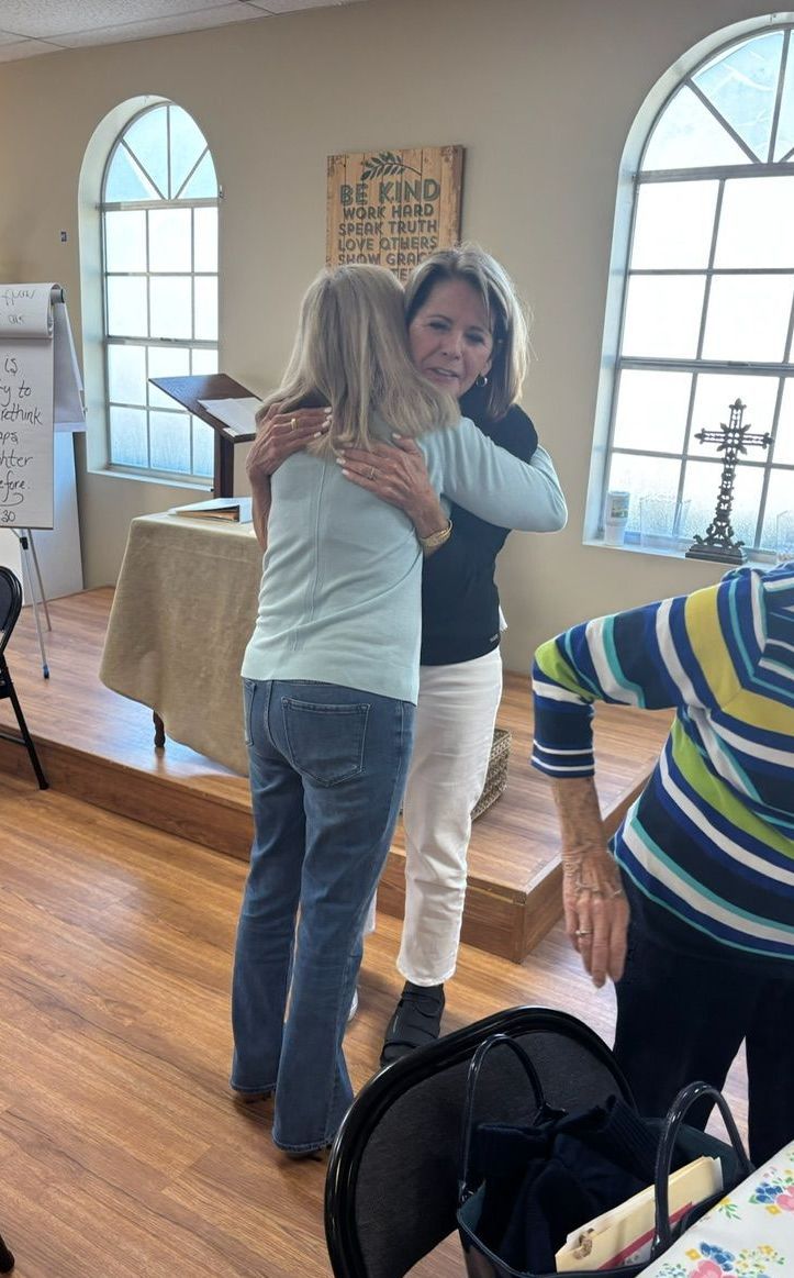 Two women embracing inside a light-filled room with wooden floors, a podium, and arched windows.