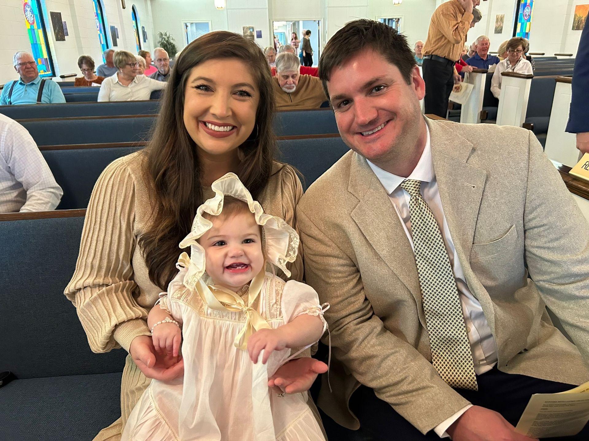 Family at church: smiling parents with baby in a christening gown; seated in pews.