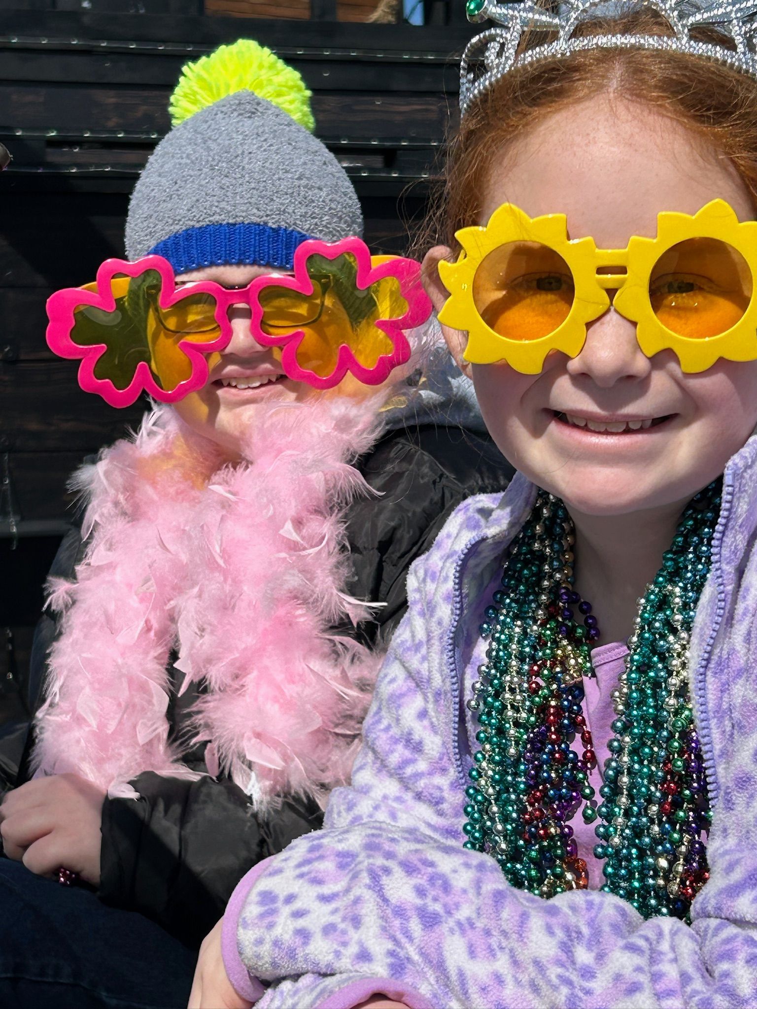 Two smiling girls wearing fun sunglasses, hats, and beads. One has a boa, the other a tiara.