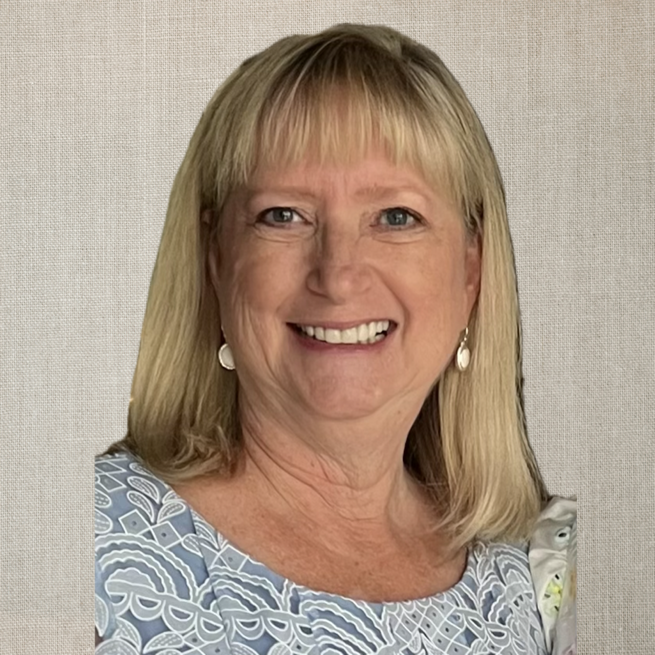 Blonde woman with a pleasant smile, wearing a patterned blue top, standing in front of a neutral backdrop.