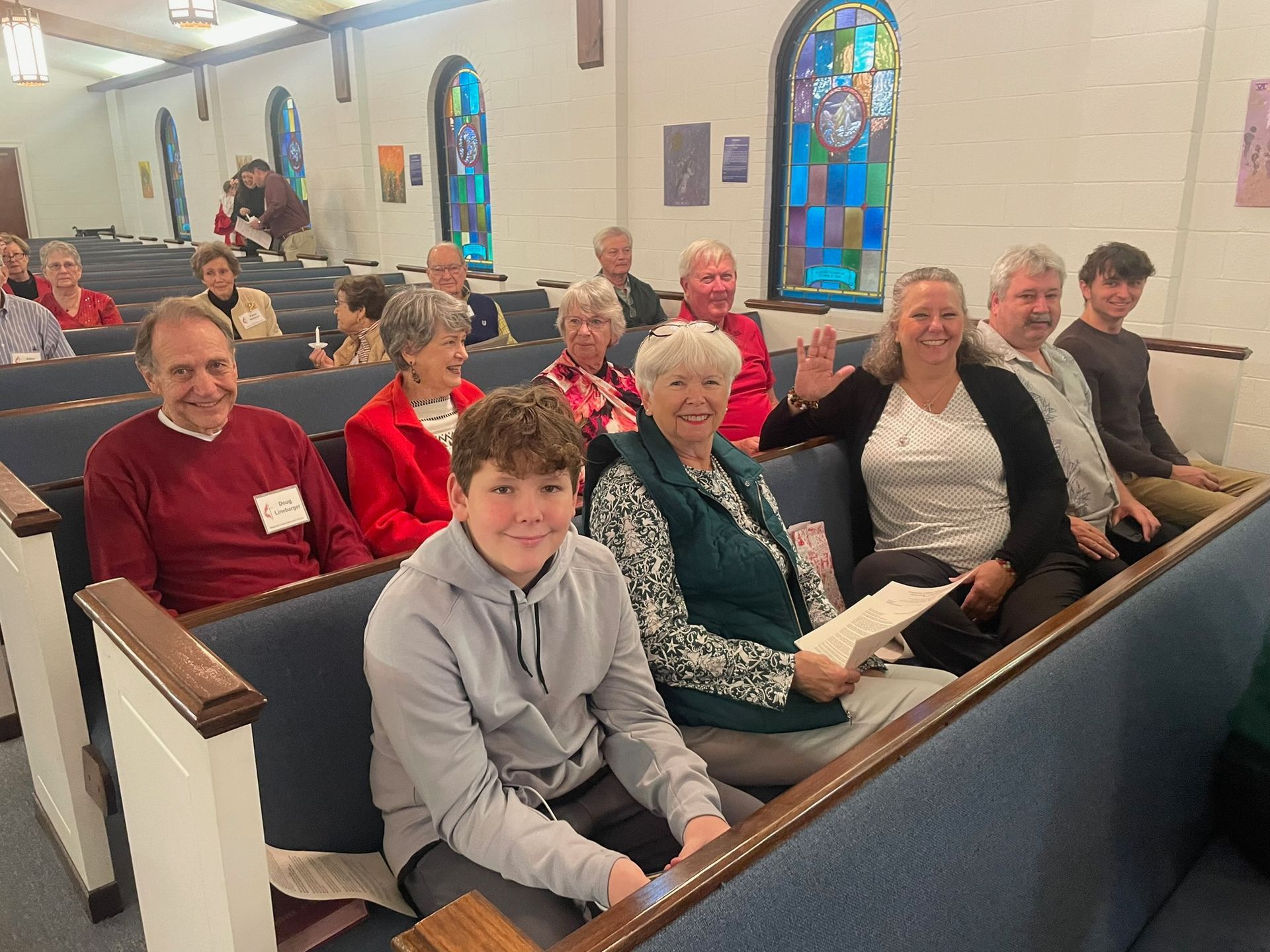 People seated in church pews. Some are smiling, and a woman waves. Sunlight streams through stained glass windows.