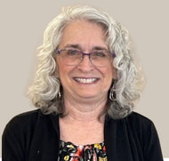Woman with gray curly hair and glasses smiles, wearing a black cardigan over a floral top.