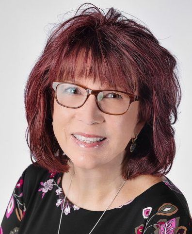 Woman with red hair, glasses, and floral top smiling at the camera.