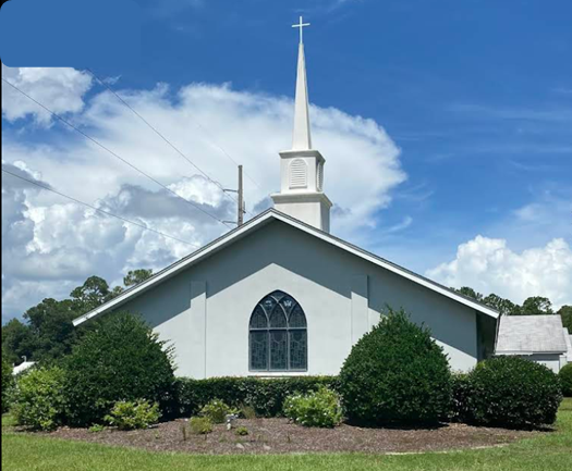 Church with a steeple and cross against a cloudy blue sky, surrounded by green bushes and grass.