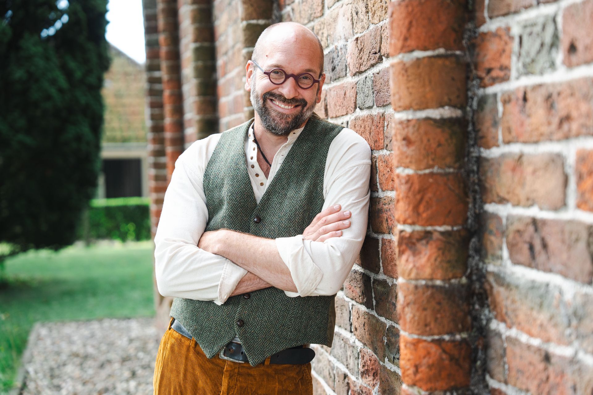 Bas leunt glimlachend tegen een bakstenen pilaar. Hij draagt ​​een vest, een wit overhemd en een gouden broek.