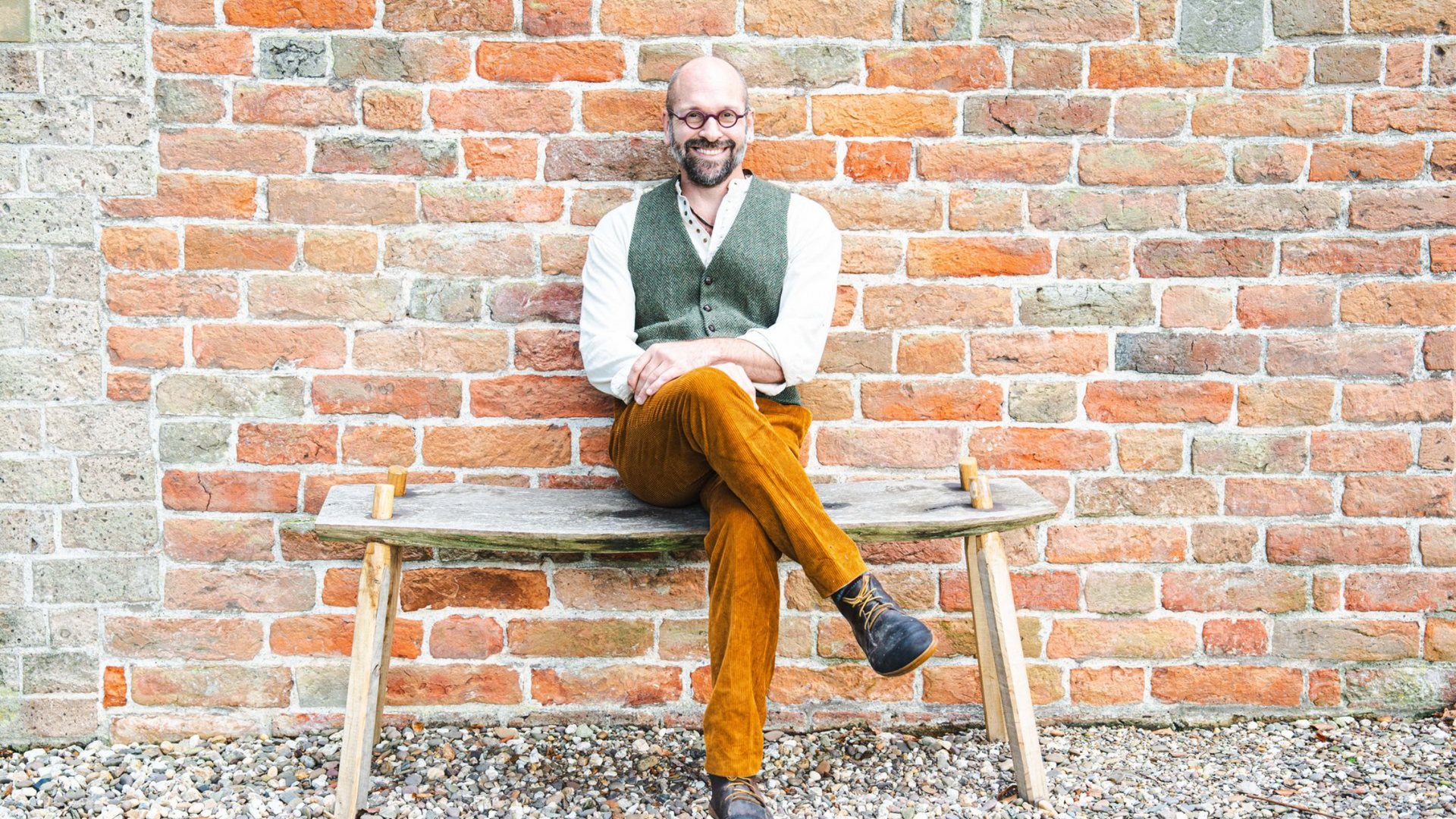 Een man in een vest en een gele broek zit op een verweerde houten bank voor een bakstenen muur.