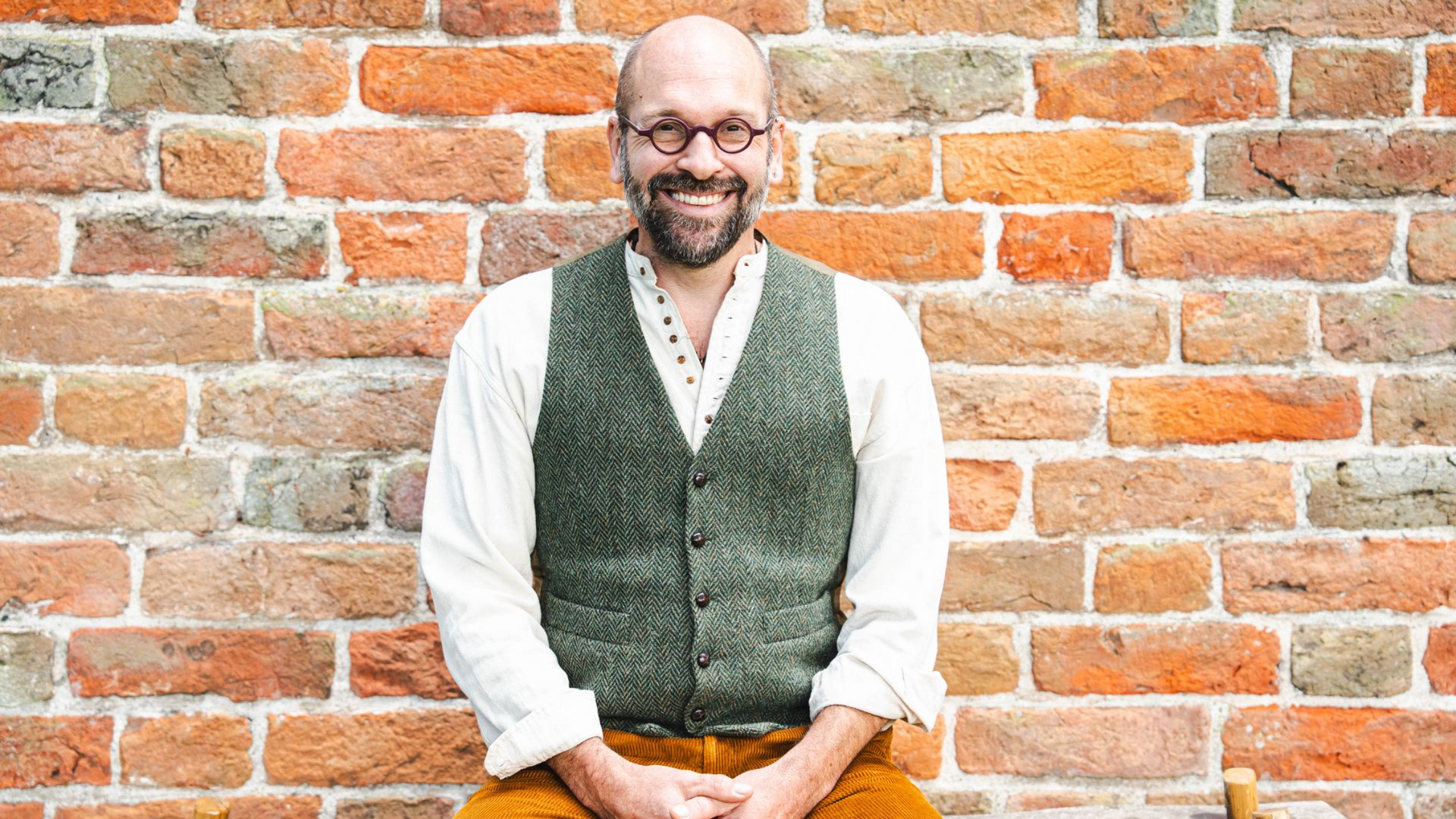 Bas, gekleed in een vest, zit voor een bakstenen muur.