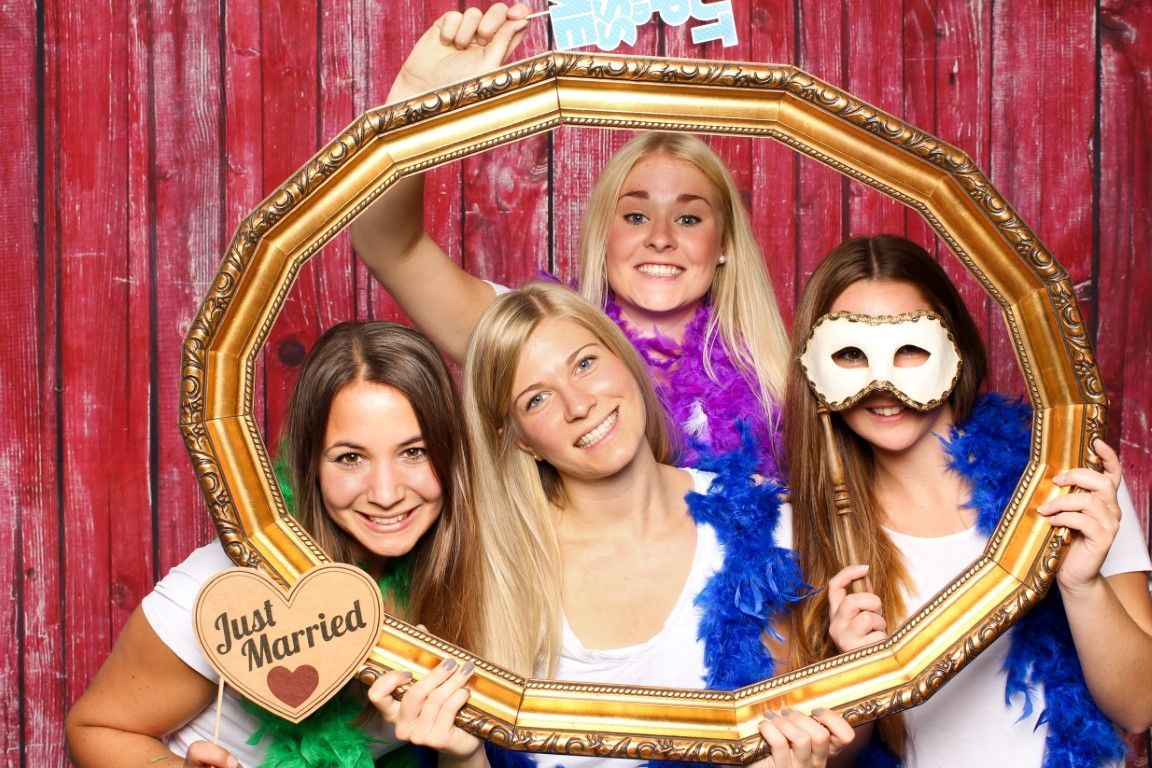 Four women posing inside a gold frame, one with a mask, holding props, smiling. Red wood backdrop.