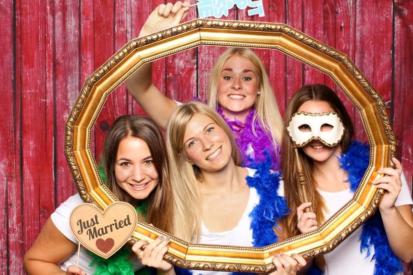 Four women posing inside a gold frame, one with a mask, holding props, smiling. Red wood backdrop.