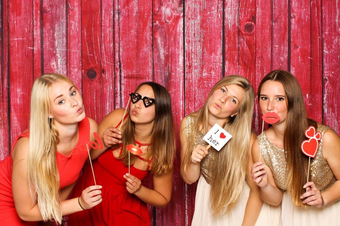 Four young women pose with props in a photo booth; red wooden backdrop.