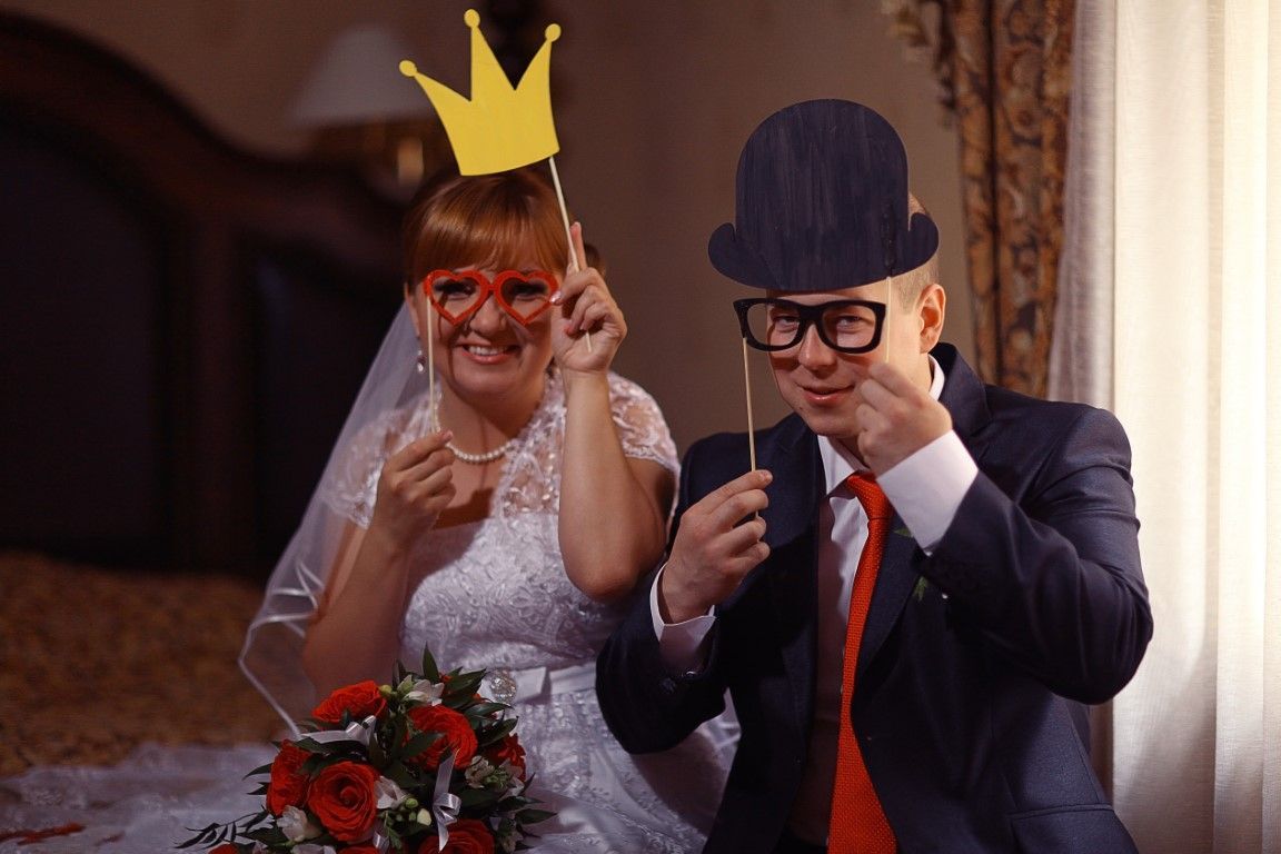 Bride and groom with silly props, smiling. Bride holds a crown, groom a top hat and glasses.